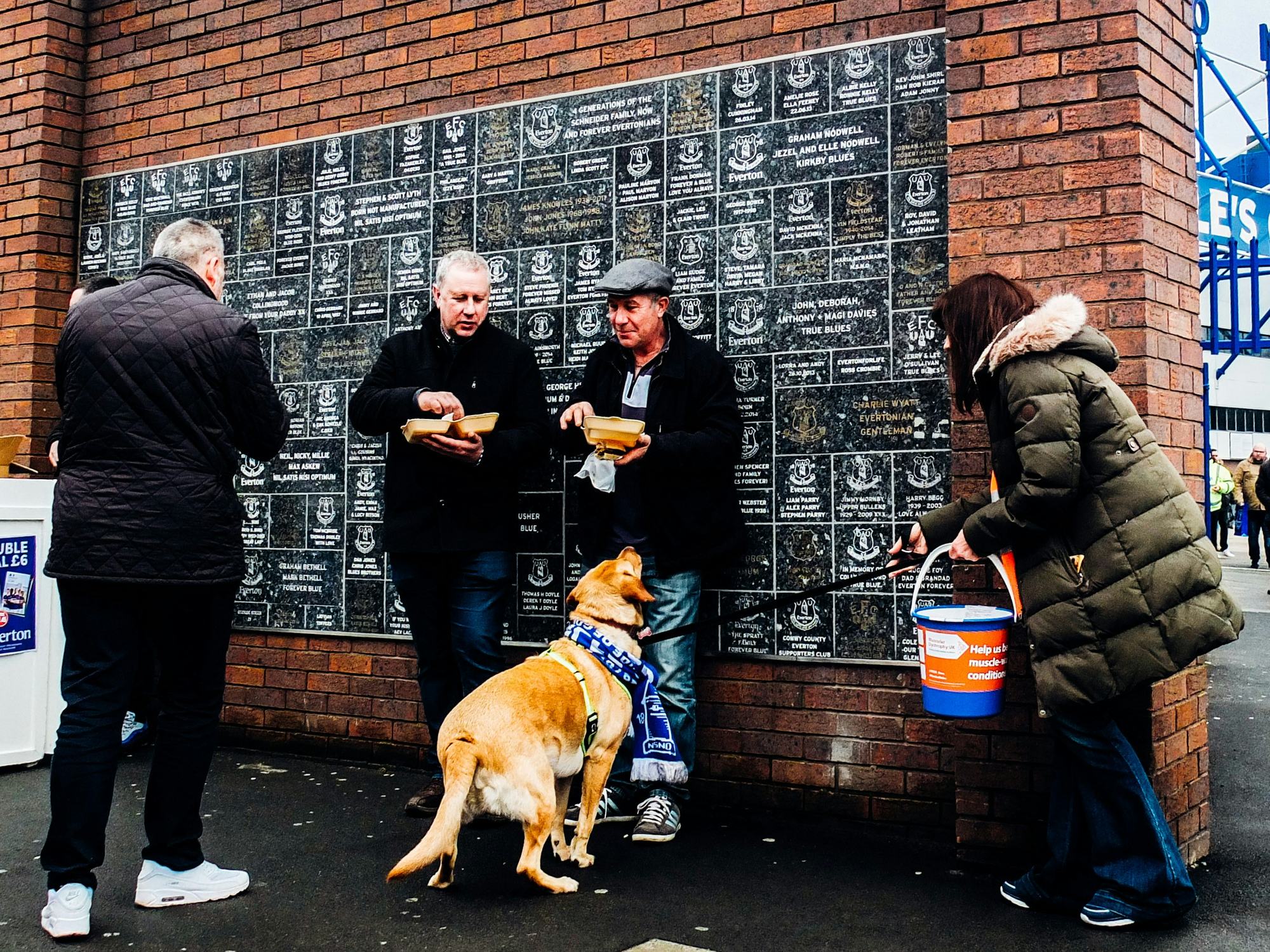 Supporters and a dog in a club scarf beside a memorial brick wall outside a ground